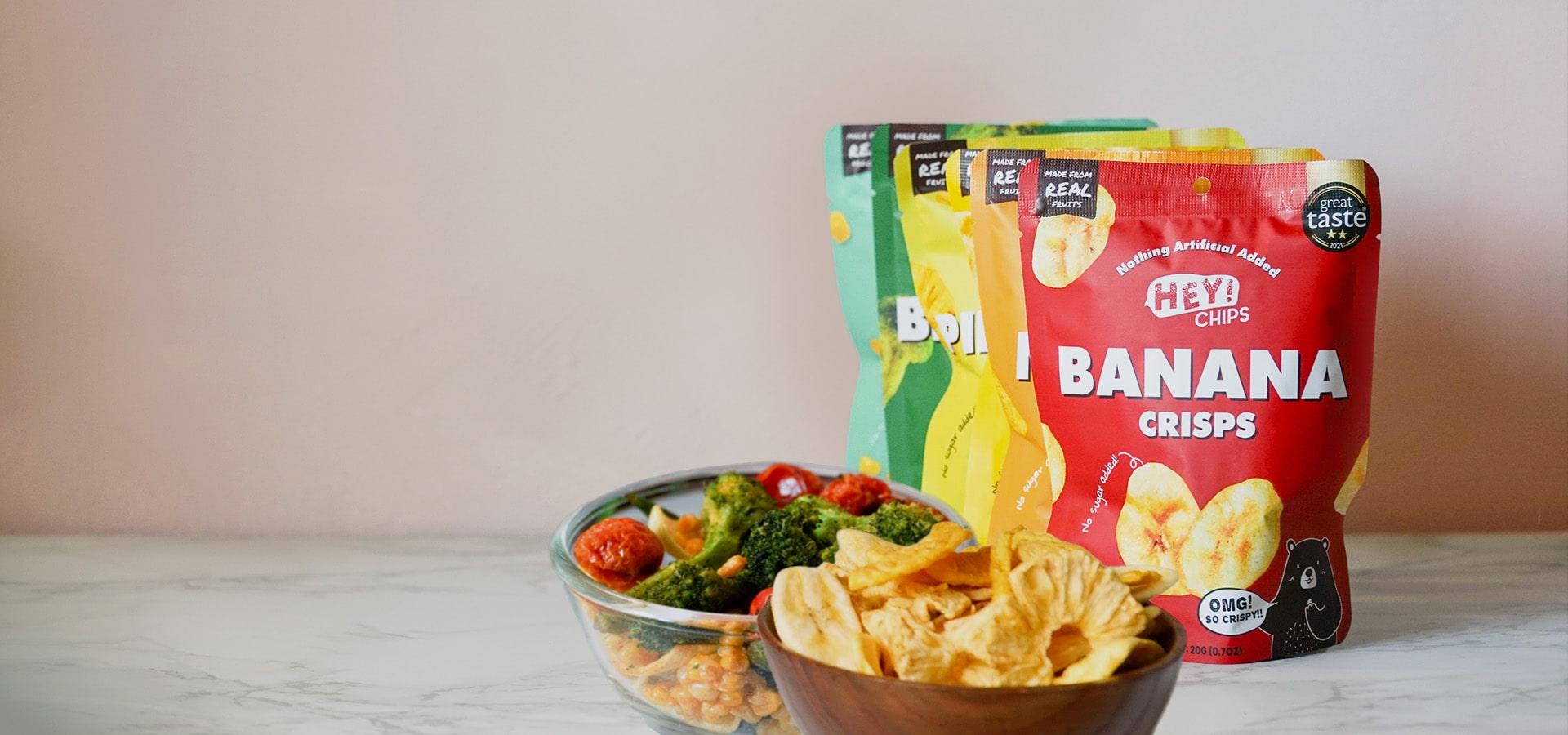 Hey! Chips pineapple and broccoli crisps packs with snacks served in a glass and wooden bowl.
